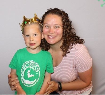 A woman and a young boy smile at the camera. The boy wears a crown and green shirt, the woman wears a pink striped shirt.