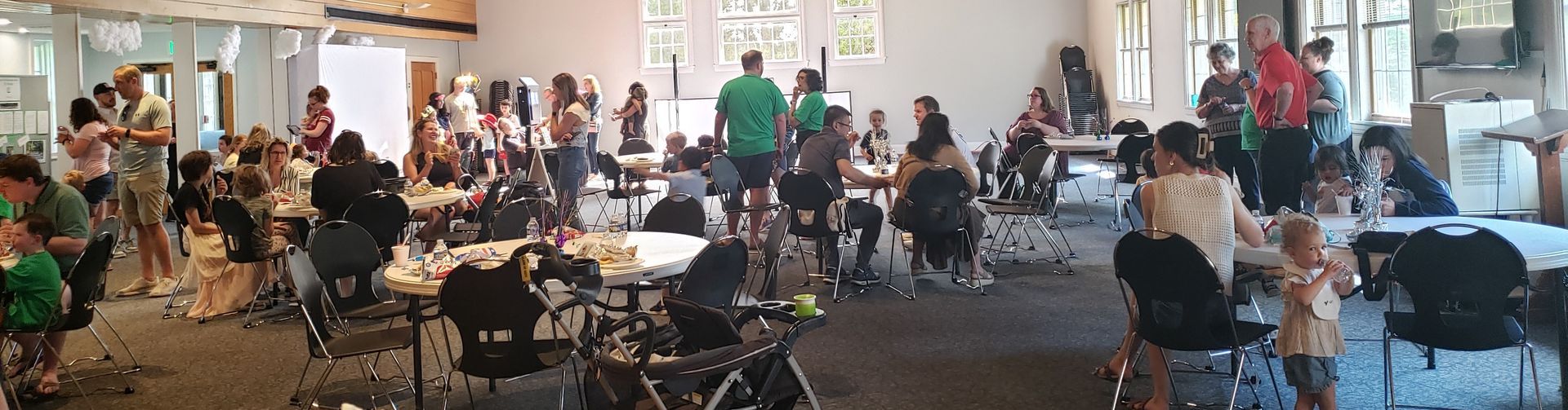 A crowded room of people at round tables, some wearing green. The room has large windows, and some individuals are standing.