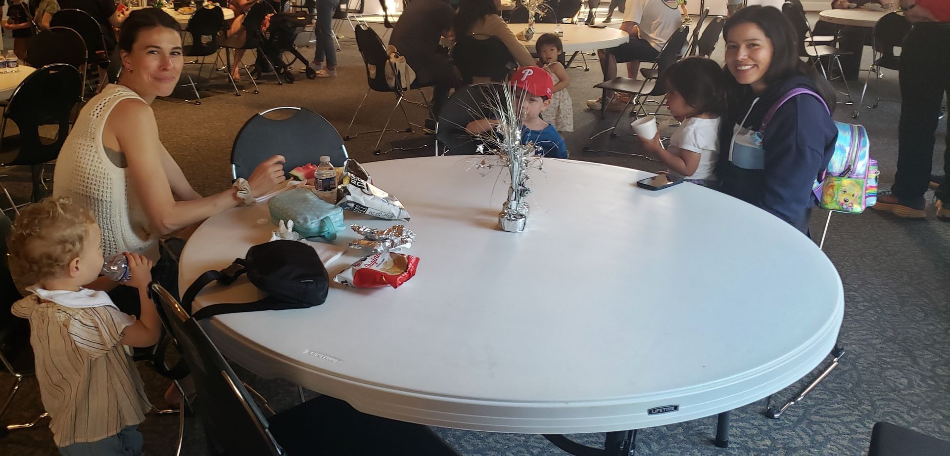 Two women and two young children sit around a large round table in a public space, likely a cafe or event venue.