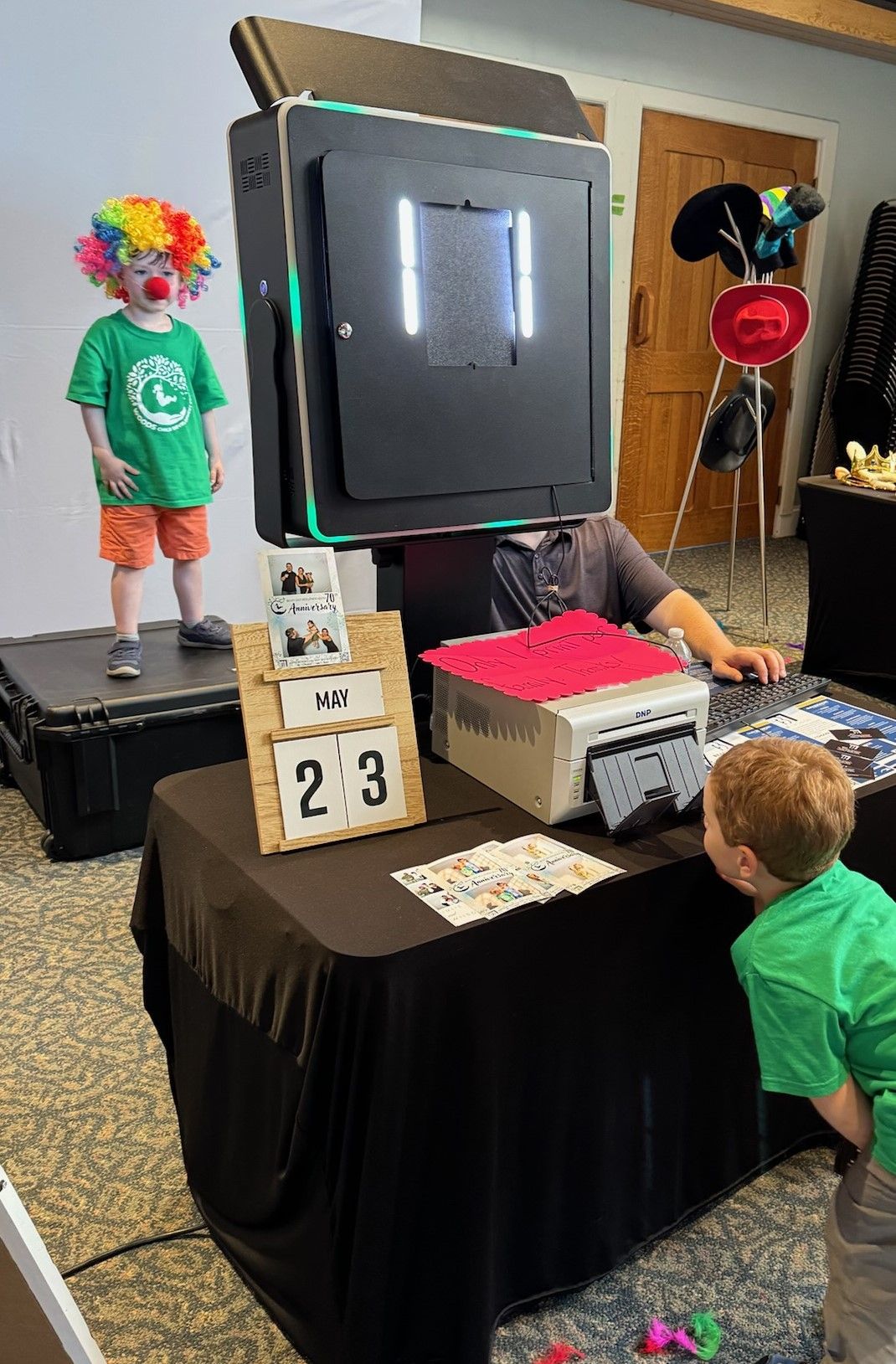 Photo booth with a clown-dressed child posing, a man operating it, and a boy looking at the prints. A printer and a number display are on the table.