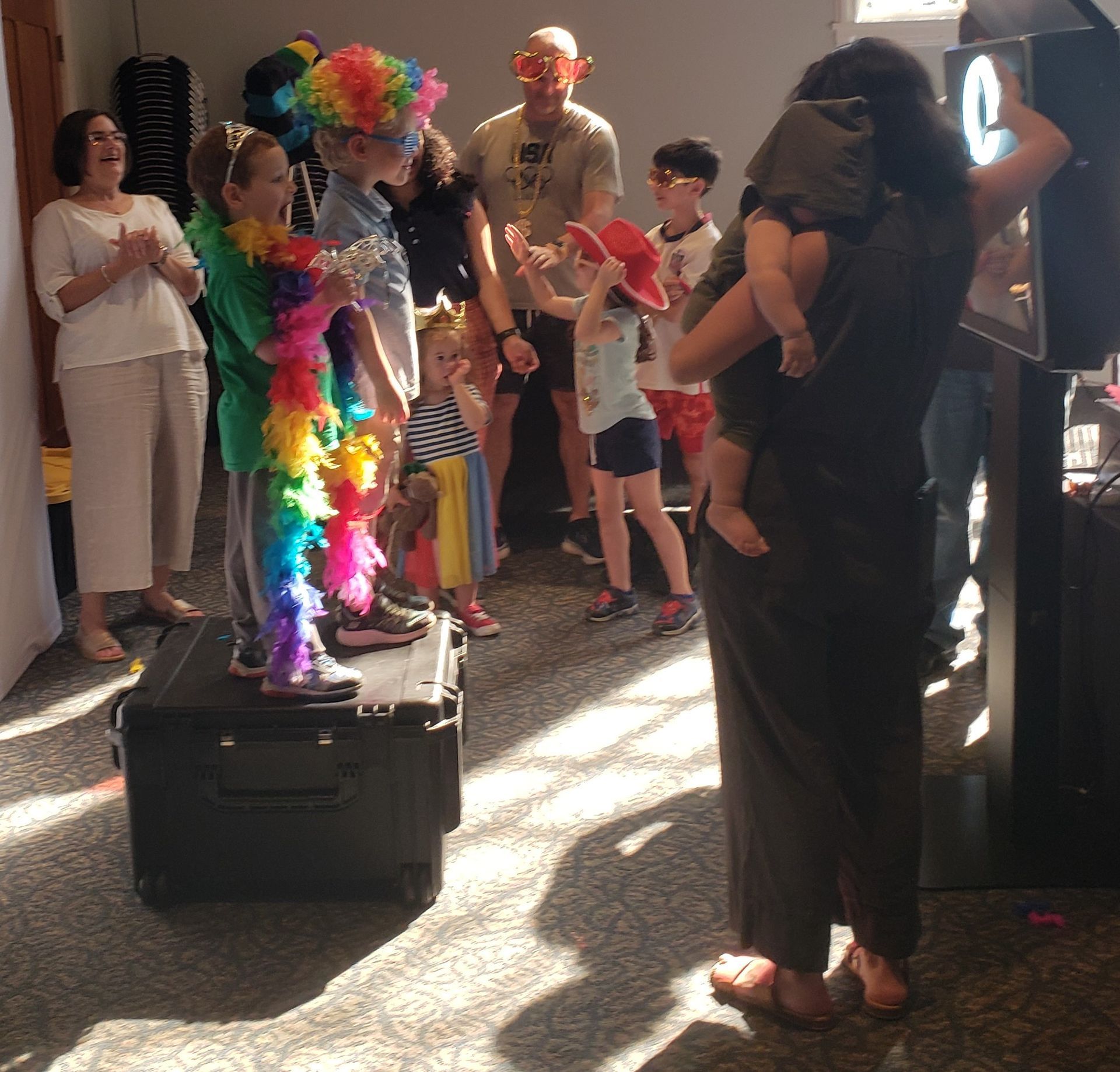 A group of people posing for a photo booth. Kids wear costumes, while a woman holds a baby. Sunlight streams in.