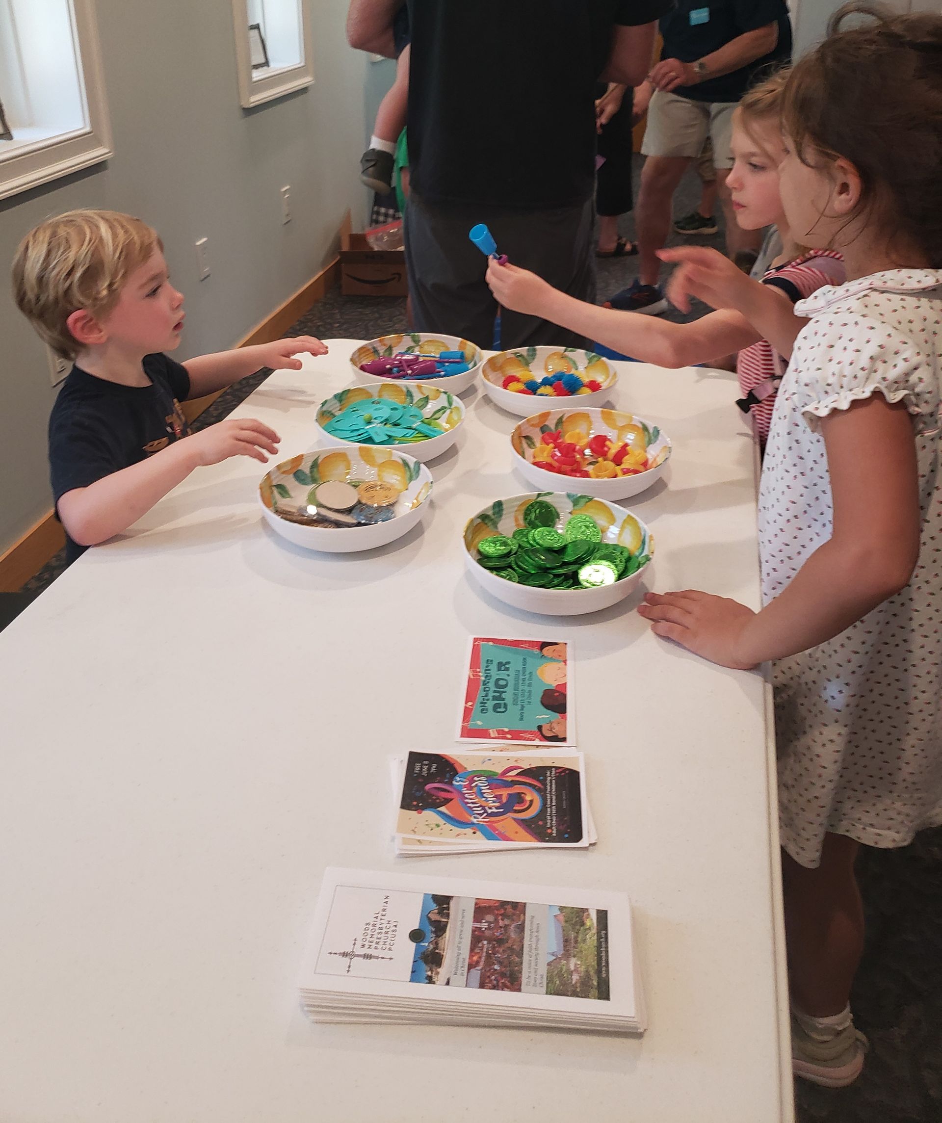 Children at a table with bowls of colorful craft supplies. Books and adults are in the background.