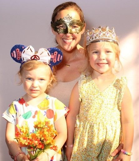 Woman and two young girls, all smiling, wearing masks and crowns. The girls hold flowers.