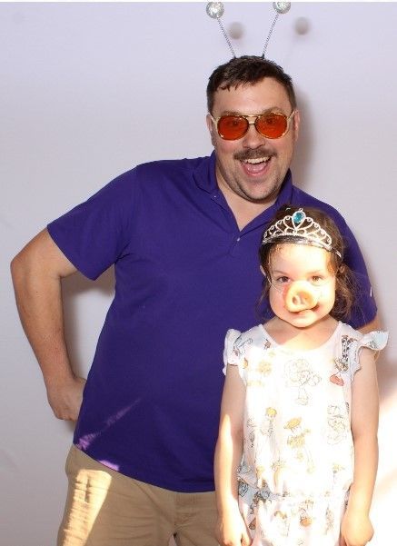 Man with orange sunglasses and novelty antennae stands next to a young girl with a pig nose and tiara. Both are smiling.