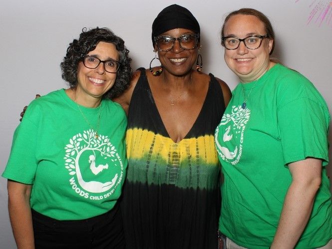 Three women smiling for a photo. Two wear green t-shirts with a logo, the other a black dress with a yellow gradient. All have glasses.