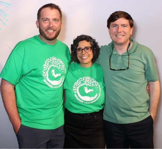 Three people smiling, wearing green shirts. Two are side by side with a woman in the middle, all posing for a photo in front of a white wall.