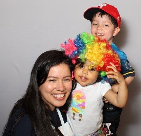 Woman smiles with a child in a rainbow wig and a boy in a baseball cap. They're all smiling at the camera.