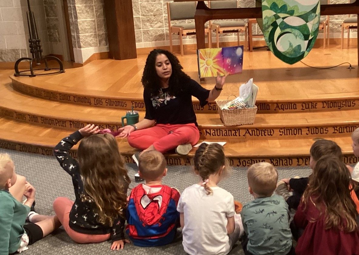A woman reads a book to a group of children sitting on a carpeted area. She is on a stage in a church.
