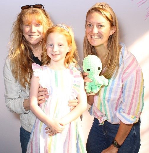 Three females smiling, posing for a photo. A young girl in a dress stands between two women, one holding a green octopus toy.
