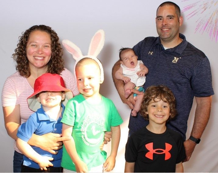 Family of six poses together; parents smile, children wear hats, one wears bunny ears, and baby is held by father.
