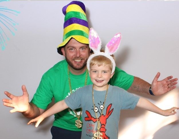 Man in green shirt and Mardi Gras hat, and a child in a lobster shirt and bunny ears, posing with arms outstretched in front of a white backdrop.