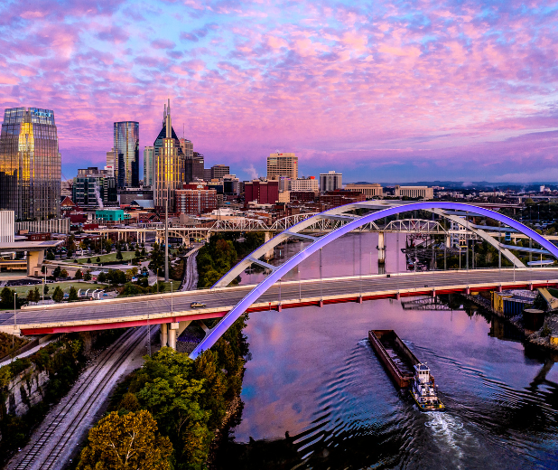 City skyline at sunset with an arched bridge over a river and a boat below, under pink clouds
