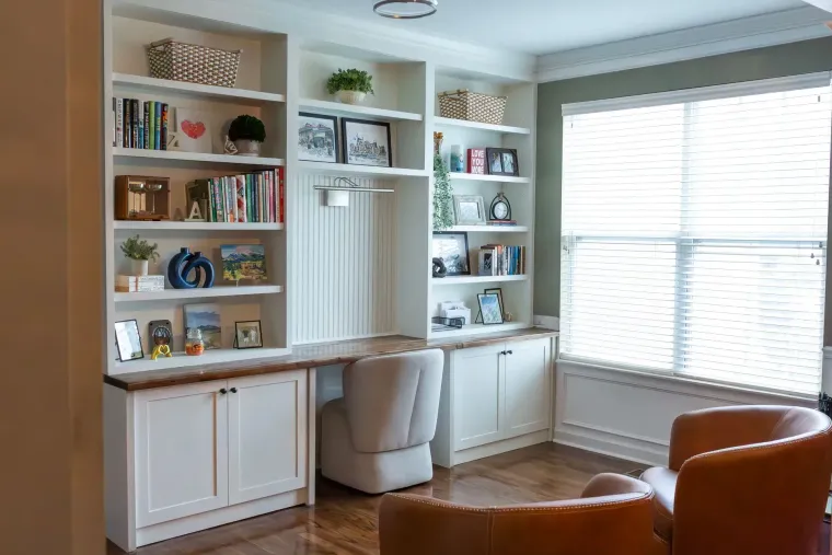 Built-in white office shelves and desk beside a bright window, with books and decor displayed.