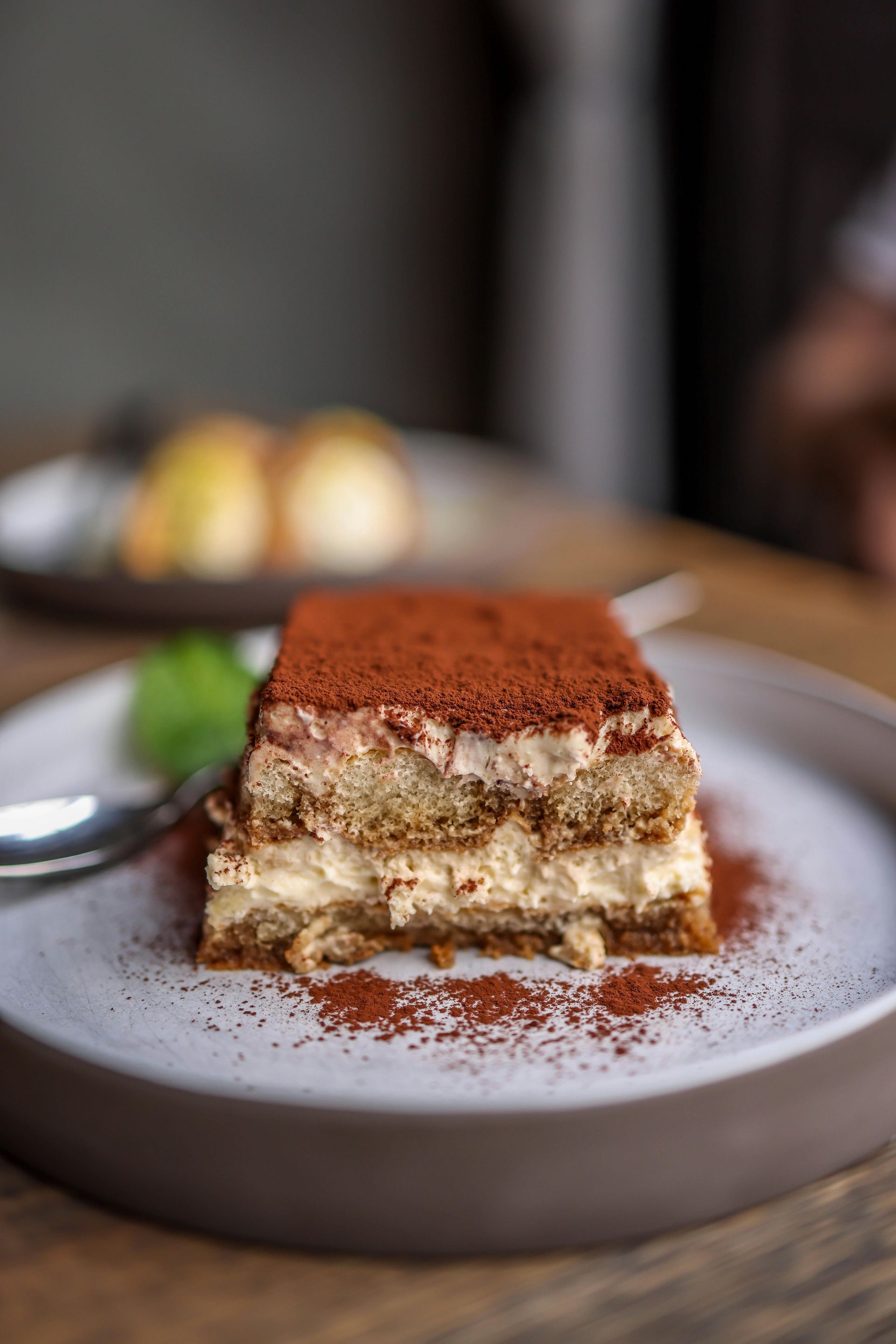 A slice of layered tiramisu dusted with cocoa powder, centered on a ceramic plate with a silver spoon and mint garnish.