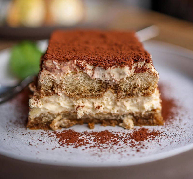 A slice of layered tiramisu topped with cocoa powder, centered on a white plate with cocoa dust, a fork, and mint.