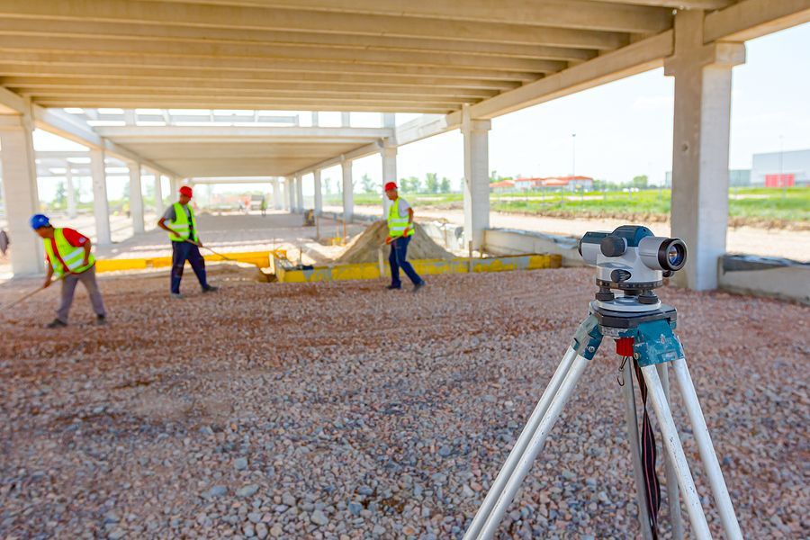 Construction workers wearing reflective vests work on a gravel ground under a canopy, with a surveying level in the front.