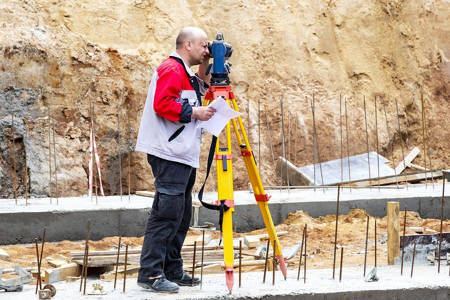 A person in a red and grey jacket uses a tripod-mounted surveying instrument on a construction site with exposed dirt.