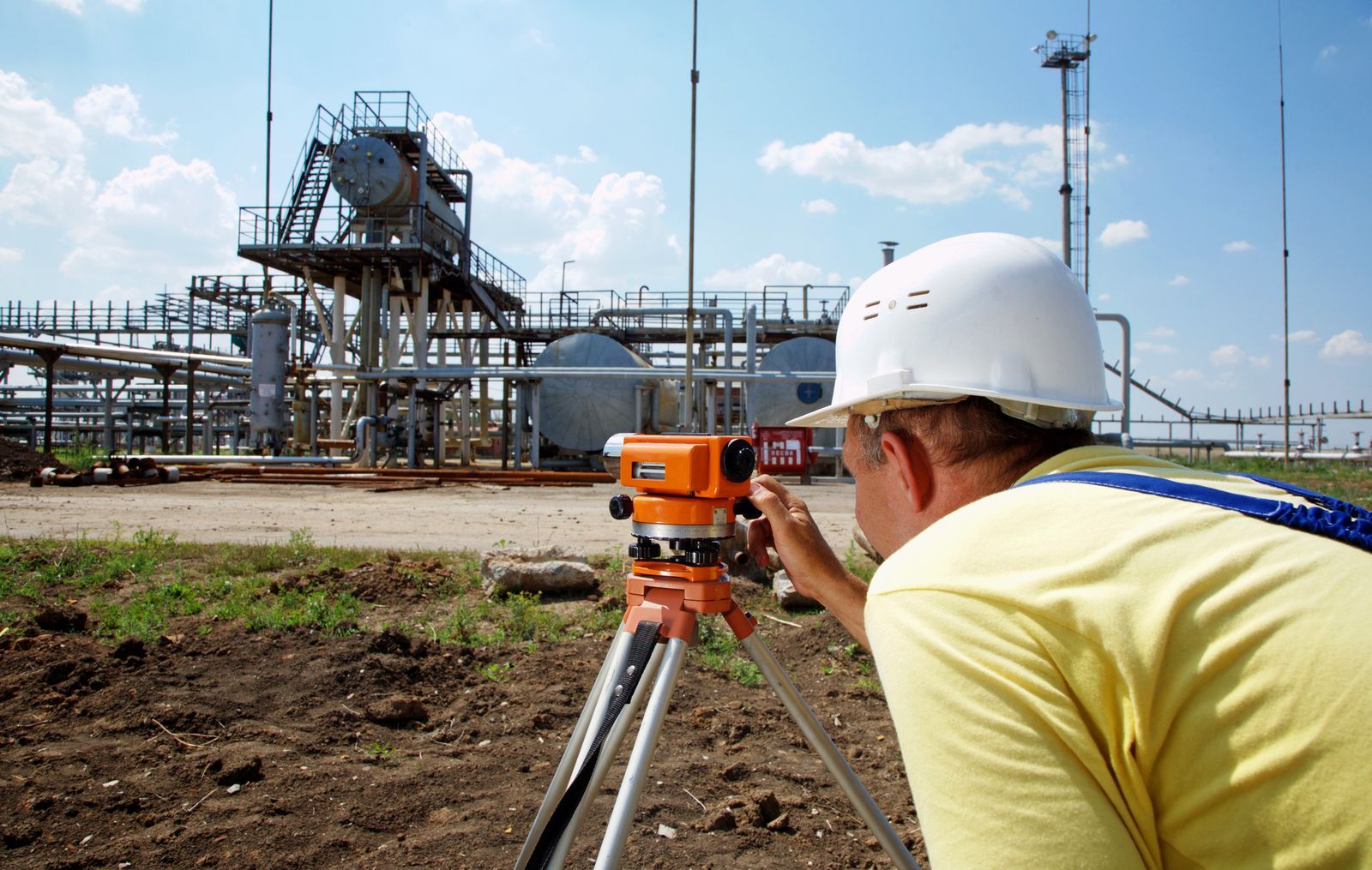 A person in a hard hat uses a surveying level on a tripod in front of an industrial oil or gas processing facility.