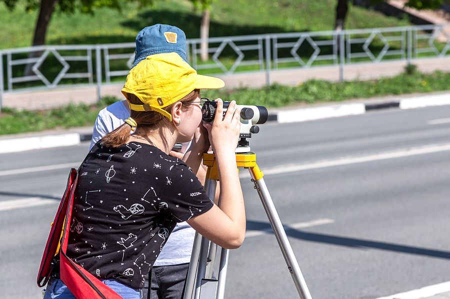 Two people stand on the edge of a road using a surveyor's level mounted on a tripod to take measurements.