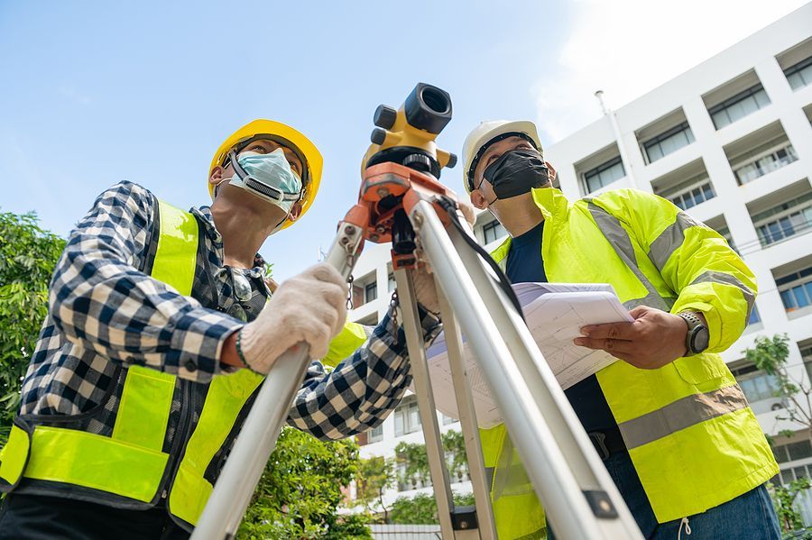 Two construction workers in high-visibility vests and helmets use a surveying instrument on a tripod outdoors.