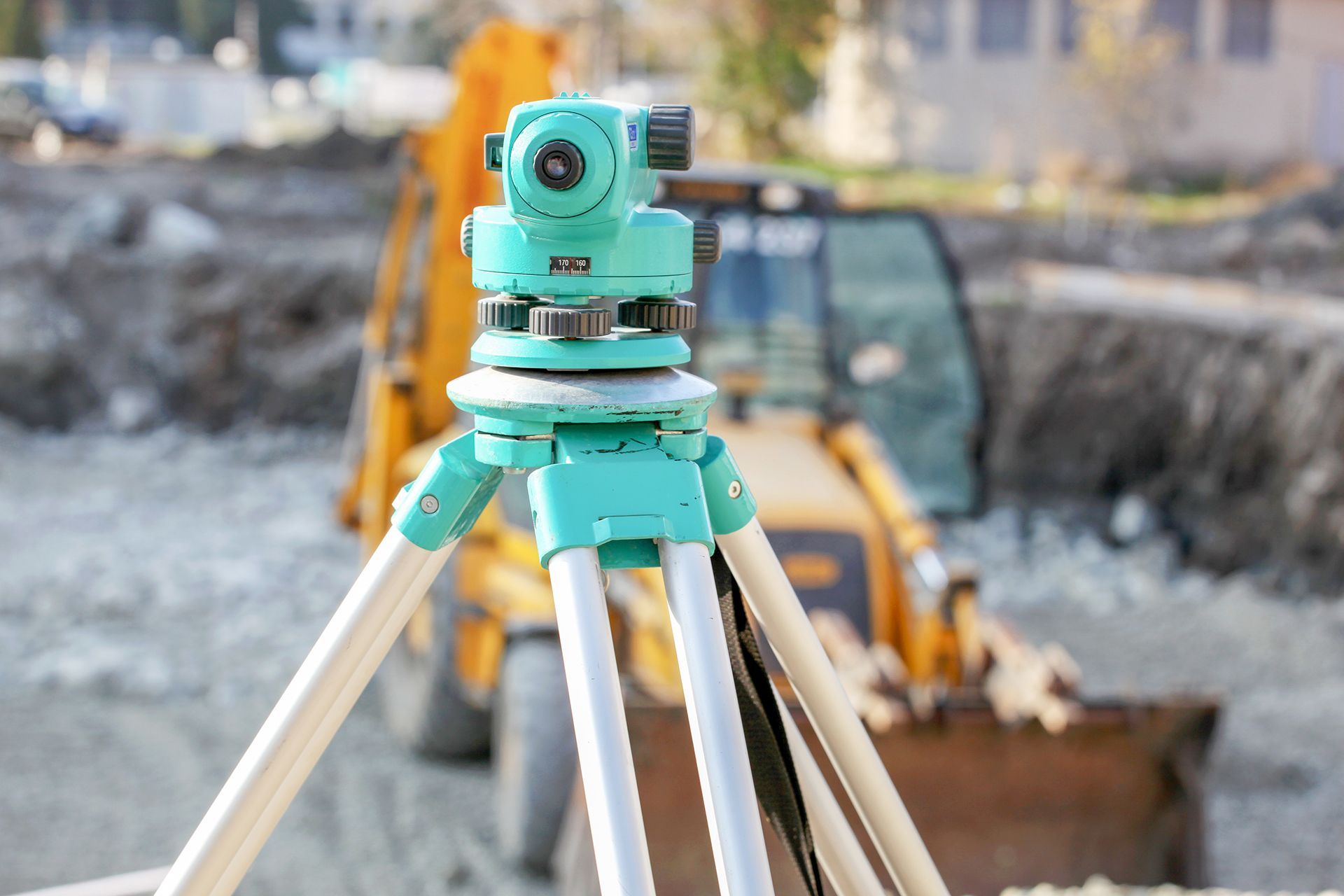 A teal surveying level mounted on a tripod at a construction site with a yellow excavator in the background.