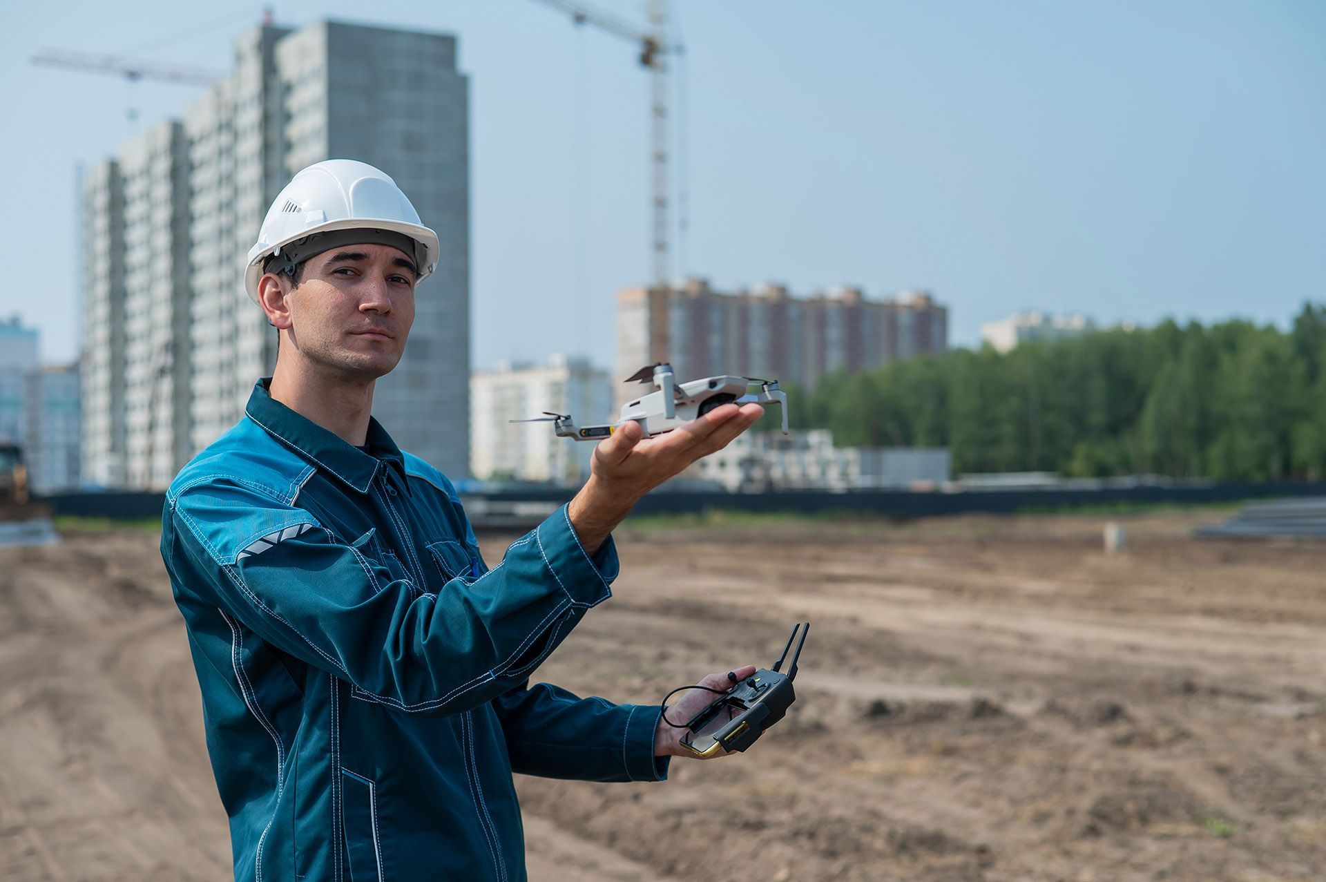 A construction worker in a white hard hat and blue uniform holds a drone while operating a controller at a site.