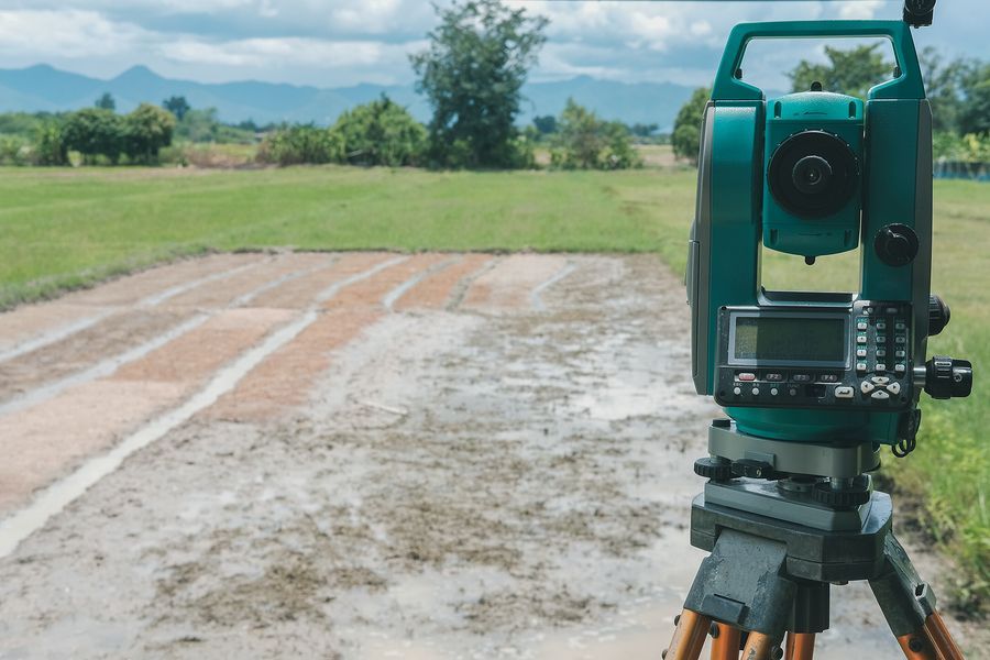 A teal surveying instrument stands on a tripod in a field with green mountains in the background.