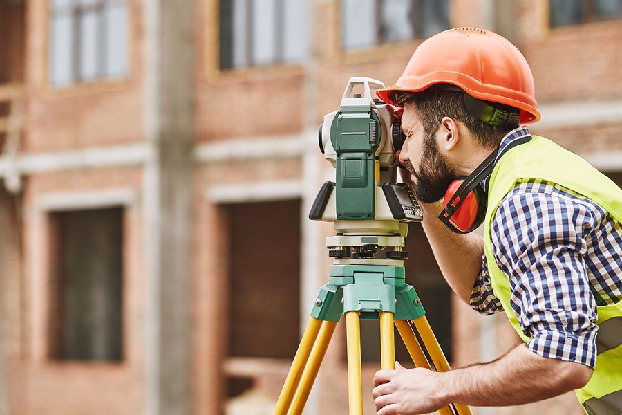 A construction worker wearing a hard hat and safety vest uses a surveying instrument on a tripod at a building site.