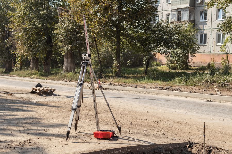 A tripod-mounted surveying instrument stands on a dirt roadside near a residential building and trees.