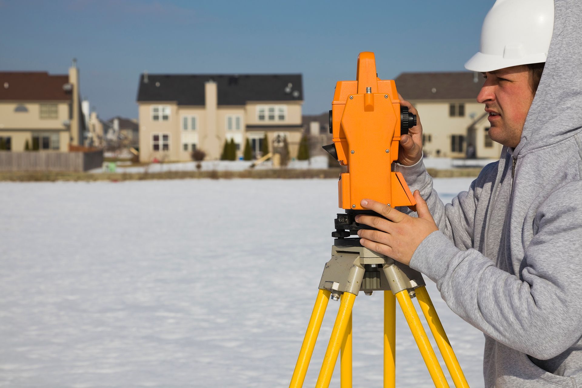 A person in a hard hat and grey hoodie uses an orange surveying instrument on a tripod in a snow-covered field.