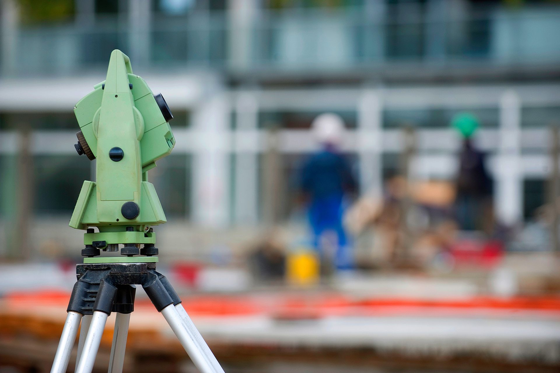 A green surveying total station on a tripod in the foreground with construction workers blurred in the background.