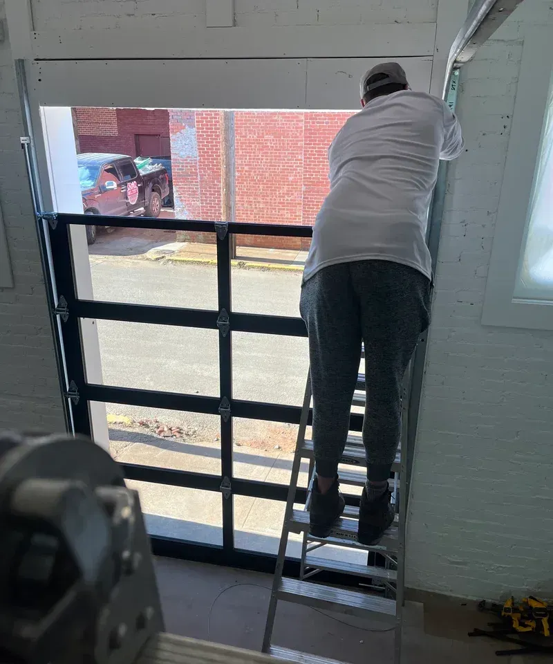Person on a ladder installing a glass garage door. Inside a building, sunny exterior visible.