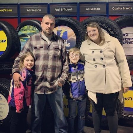Family of four posing in front of large tire display at a Goodyear store, wearing coats. | D&D Tire & Auto Center