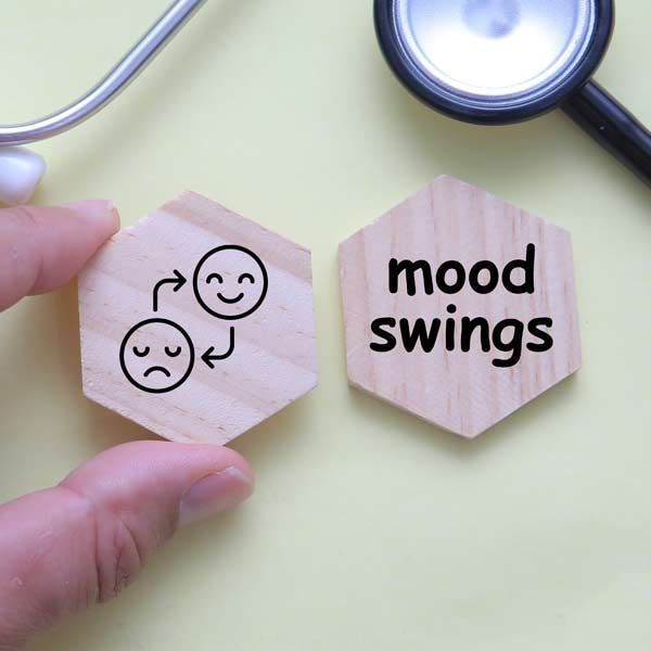 Hand holding a wooden hexagon with a sad/happy face diagram next to one that says “mood swings,” with a stethoscope in the background.