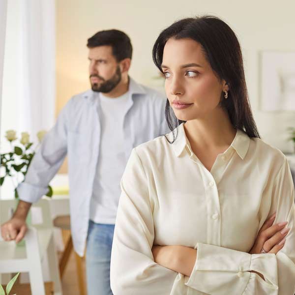 Two people standing apart in a bright room after agruement from chronic stress, one woman with folded arms in the foreground and a man in the background