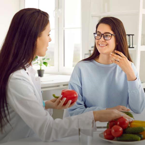 A dietitian showing a red bell pepper to a smiling patient. A bowl of vegetables on the table.