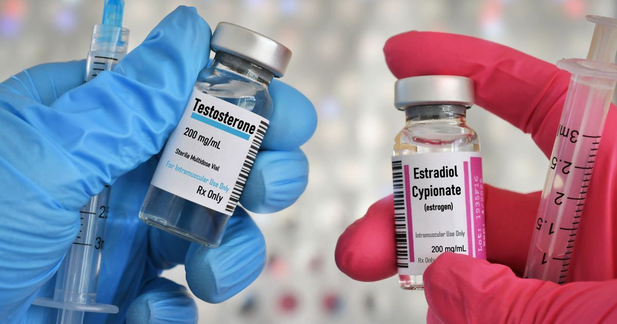 Hands wearing blue and pink gloves hold vials of testosterone and estradiol with syringes representing Hormone Replacement Therapy