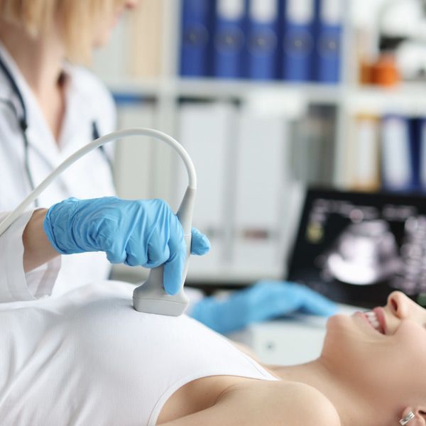 Doctor using ultrasound on a patient's chest in a medical office; blue gloves, screen displays images.