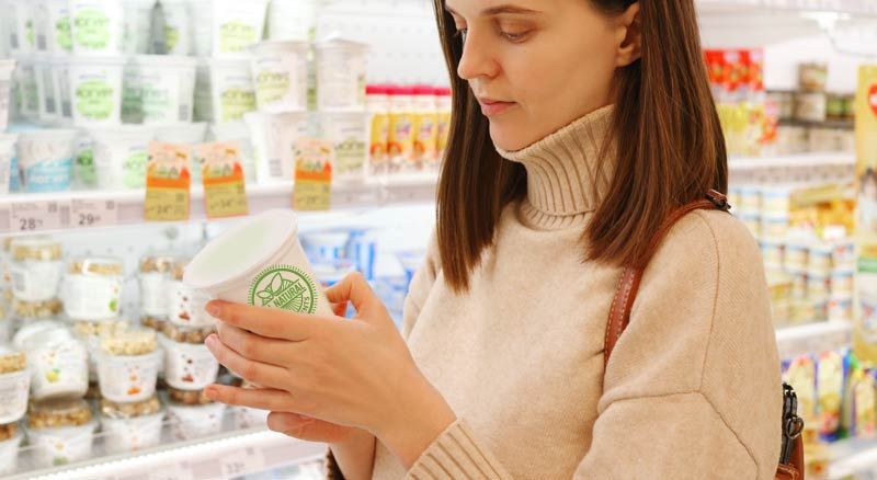 Woman in a sweater reads a container of yogurt in a grocery store.