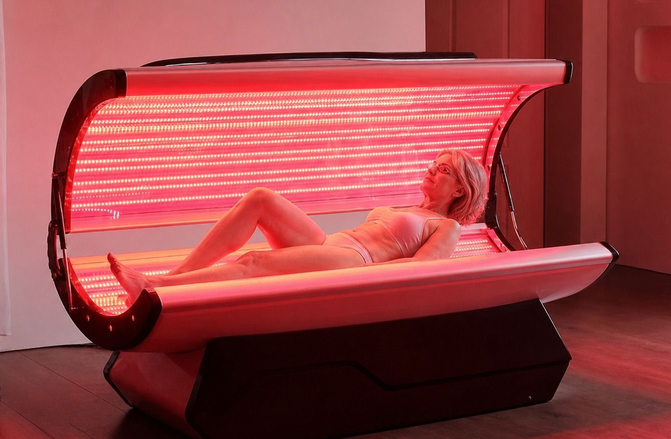 Person reclining in a red-light bed, glowing pink under the curved canopy.
