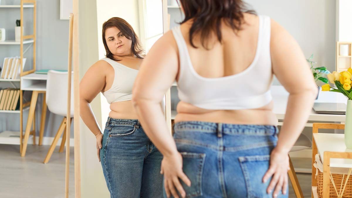 Overweight Person in a white top and jeans looking at herself in a mirror in a bright room, considering Weight Loss Resistance Treatments NYC