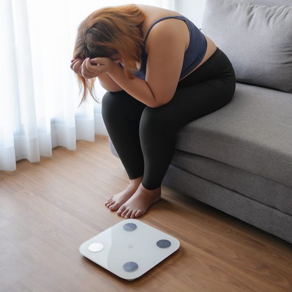 A woman struggling with weight-loss resistance sits on a couch, covering her face beside a digital bathroom scale on the floor.