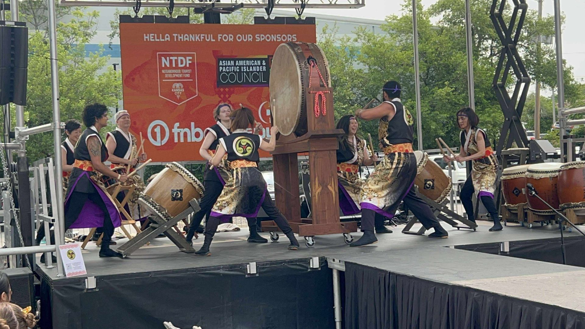 Taiko drummers perform on stage outdoors. Drummers wear black tops, gold accents, and purple skirts, playing large drums.