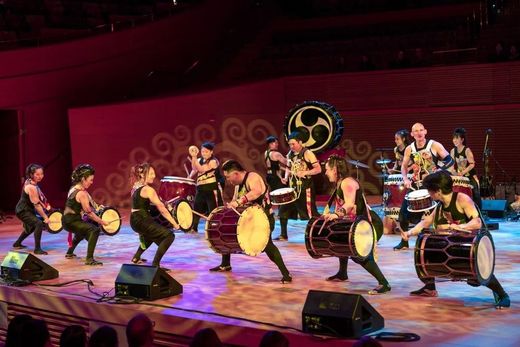 Taiko drummers perform on stage; red backdrop, drums, energetic action.
