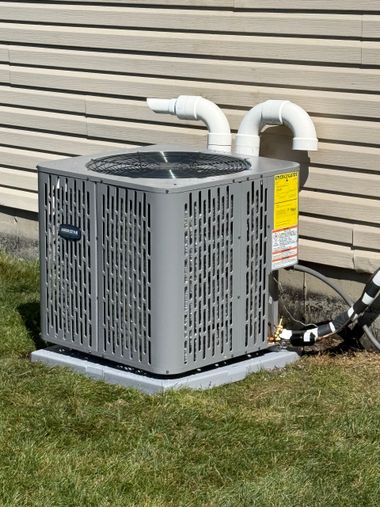 Outdoor air conditioner unit beside a house, with white pipes and a yellow warning label.