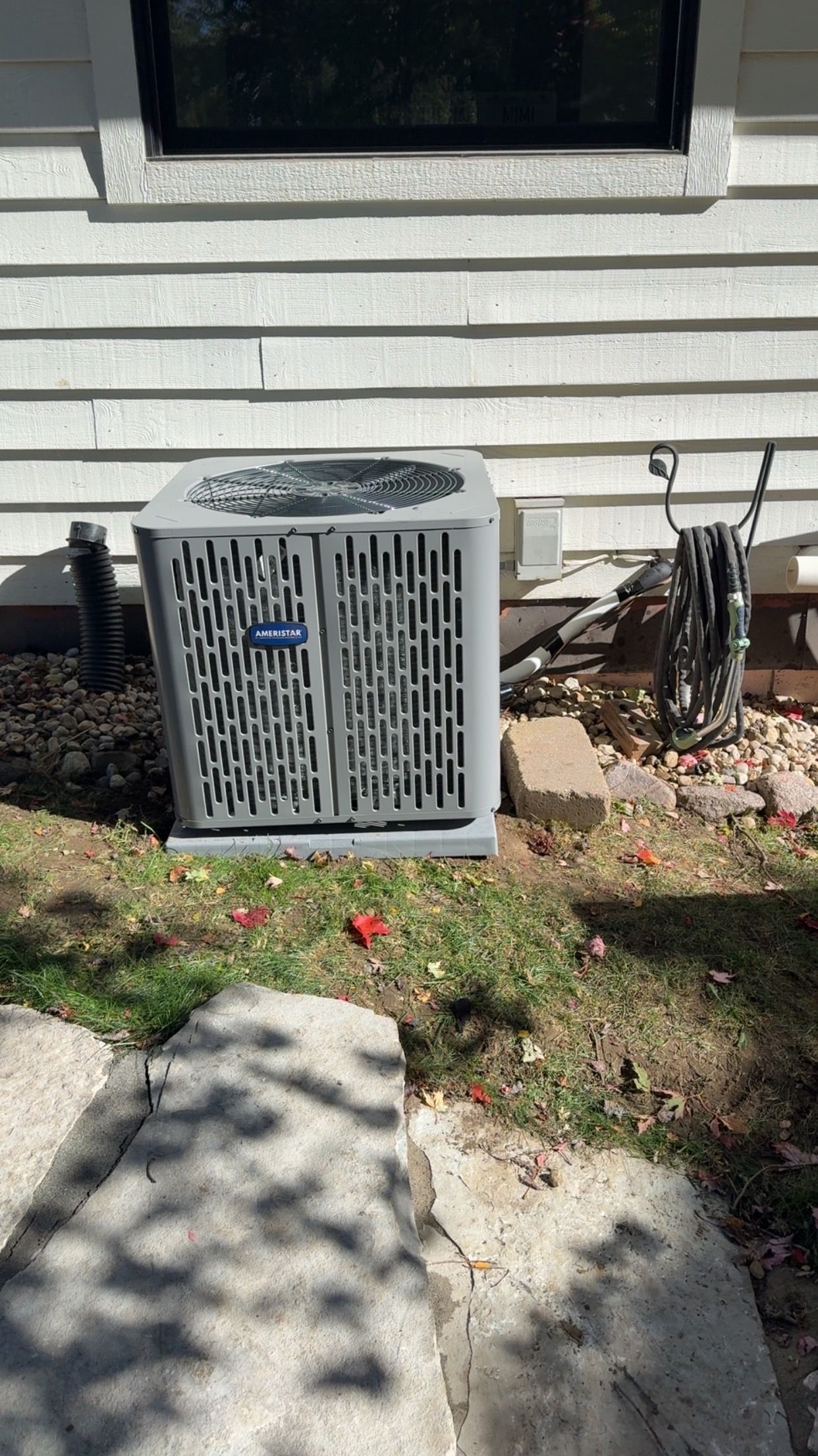 Outdoor air conditioner unit beside a house with white siding, near a garden bed and sidewalk.
