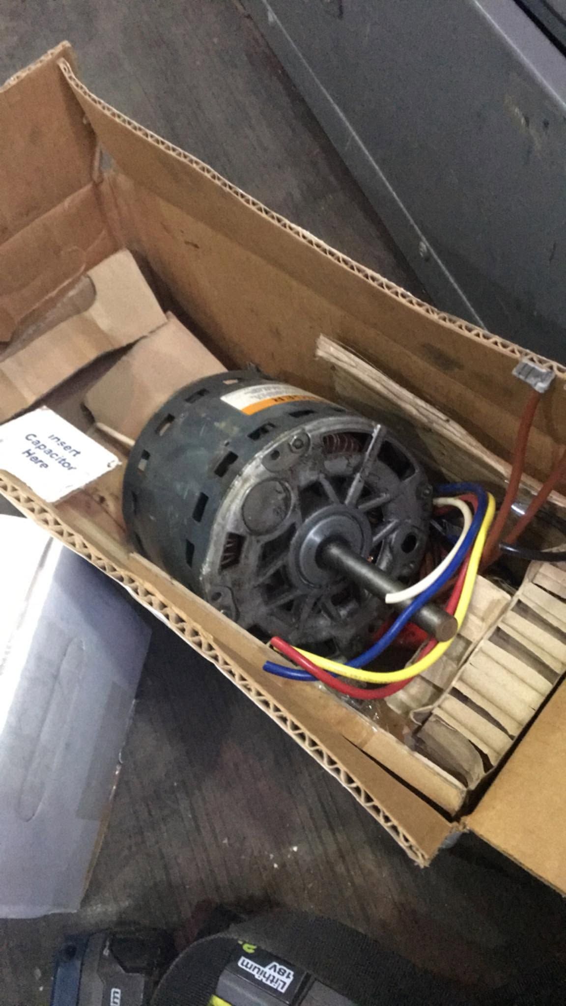 Electric motor in a cardboard box, with colorful wires attached, resting on a gray floor.