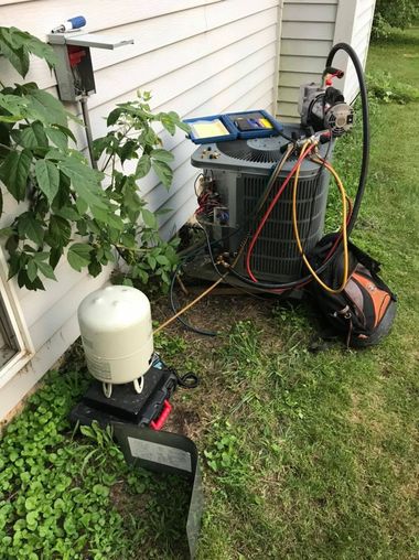 HVAC service equipment beside a house, with hoses, gauges, and a white tank on the grass.