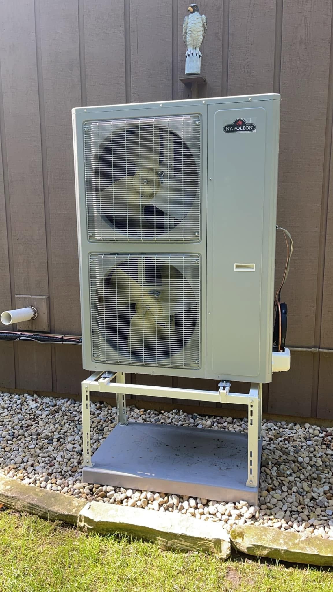 Outdoor air conditioning unit on a metal stand beside a wooden fence and gravel ground.