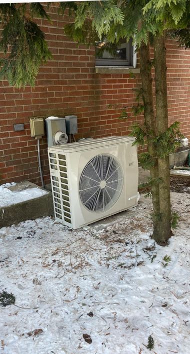 Outdoor air conditioner unit beside a brick building, partly covered in snow with a tree in front.